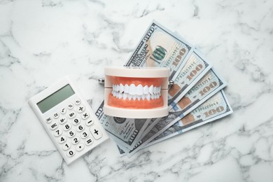 Educational dental typodont model, dollar banknotes and calculator on white marble table, flat lay. Expensive treatment