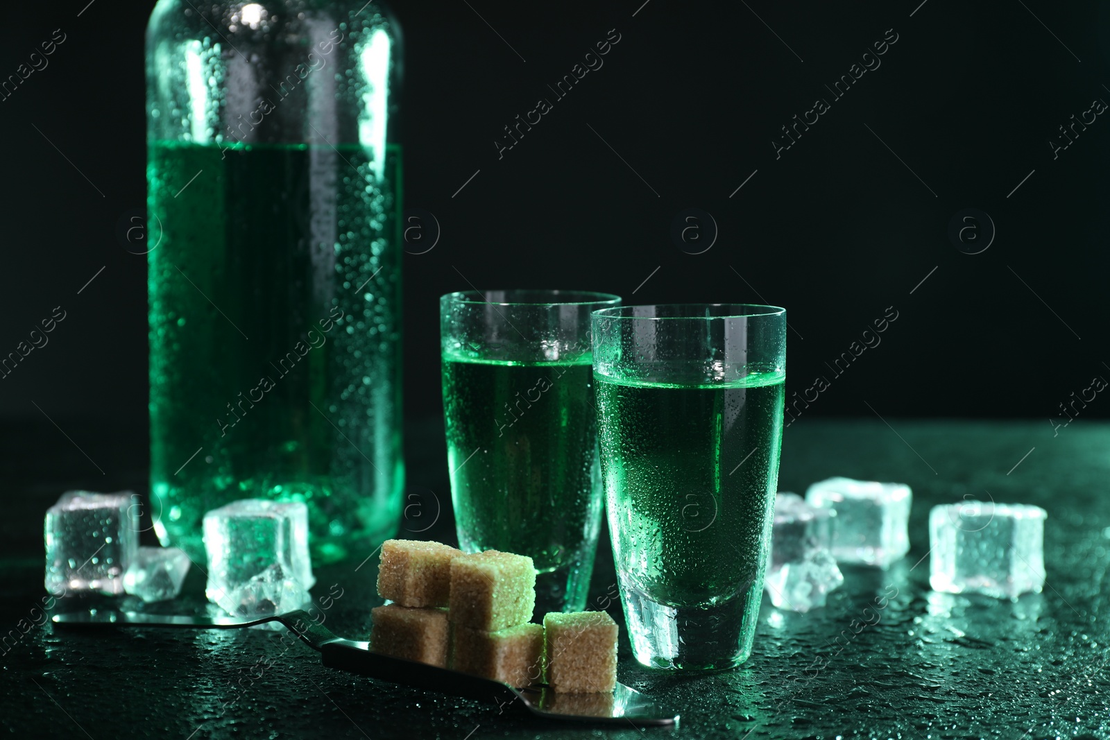 Photo of Absinthe in shot glasses, spoon, brown sugar and ice cubes on gray table against dark background. Alcoholic drink