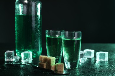 Photo of Absinthe in shot glasses, spoon, brown sugar and ice cubes on gray table against dark background. Alcoholic drink