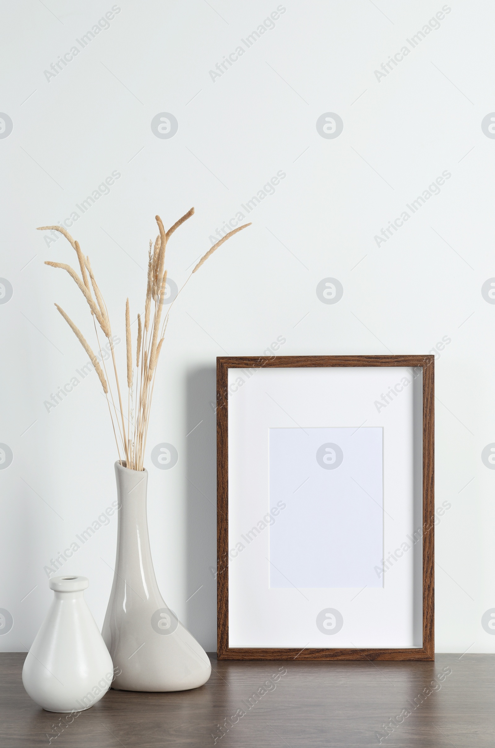 Photo of Empty photo frame and vase with dry decorative spikes on wooden table. Mockup for design