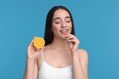 Beautiful young woman with vitamin pill and piece of orange on light blue background