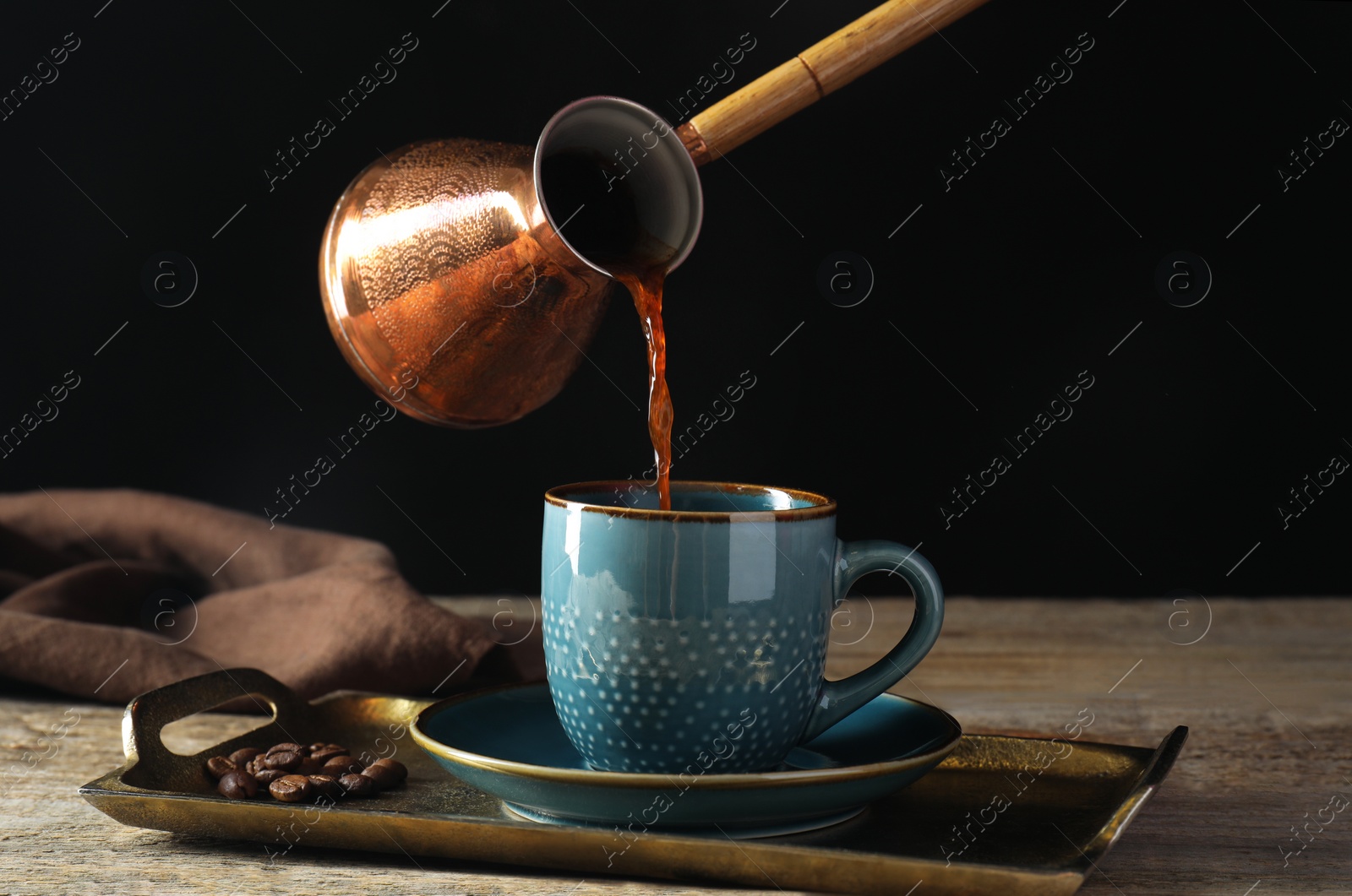 Photo of Turkish coffee. Pouring brewed beverage from cezve into cup at wooden table against black background