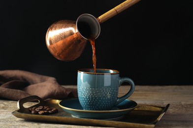 Photo of Turkish coffee. Pouring brewed beverage from cezve into cup at wooden table against black background