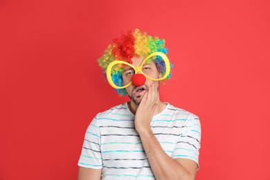 Photo of Funny man with large glasses, rainbow wig and clown nose on red background. April fool's day