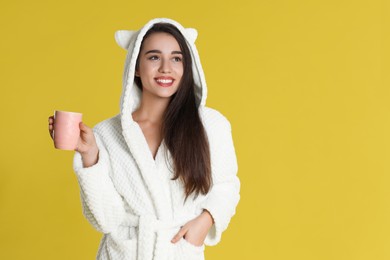 Young woman in bathrobe with cup of morning drink on yellow background. Space for text