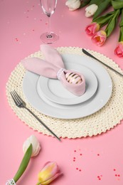 Photo of Festive table setting with painted egg, plates and tulips on pink background. Easter celebration