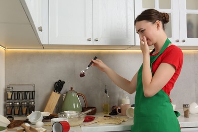 Photo of Young woman disgusted with unclean spoon in messy kitchen. Many dirty dishware and utensils on countertop