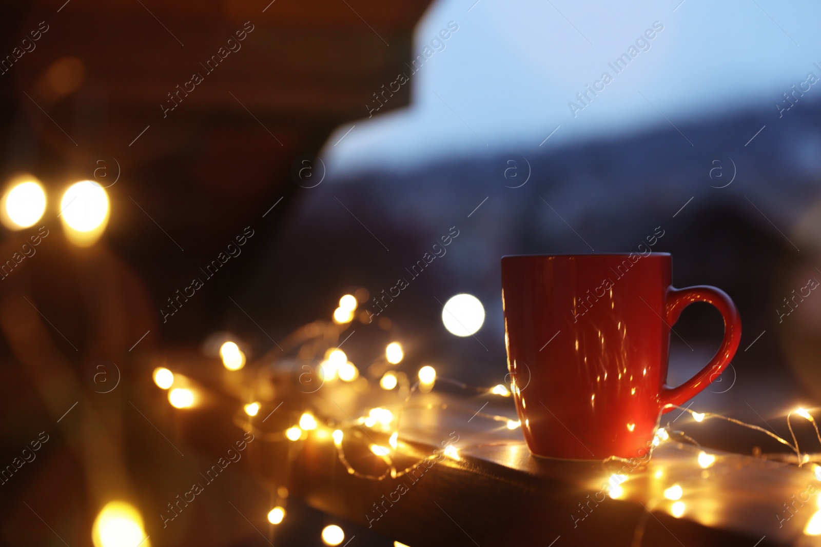 Photo of Cup of hot beverage on balcony railing decorated with Christmas lights, space for text. Winter evening