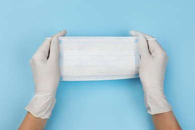 Doctor in medical gloves holding protective mask on light blue background, closeup