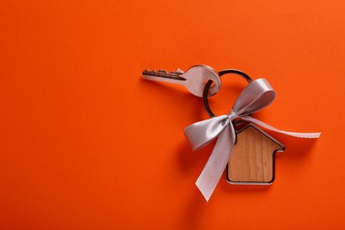 Photo of Key with trinket in shape of house and grey bow on orange background, top view. Housewarming party