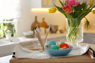 Photo of Beautifully painted Easter eggs in bowl and brushes on table indoors