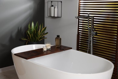 Stylish bathroom interior with ceramic tub, candles and care products on wooden bath tray