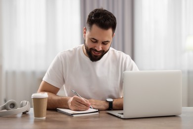 Photo of Young man writing down notes during webinar at table in room