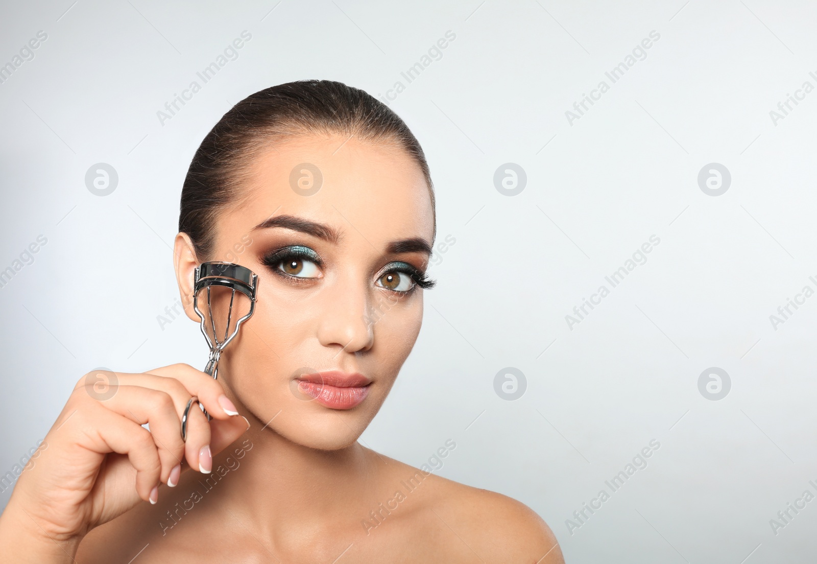 Photo of Portrait of young woman with eyelash extensions holding curler on light background. Space for text