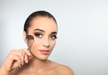 Portrait of young woman with eyelash extensions holding curler on light background. Space for text