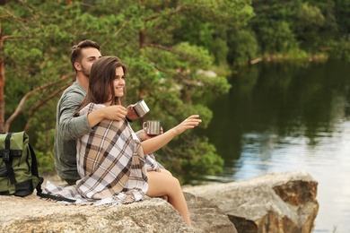 Cute couple with mugs and plaid near lake. Camping season