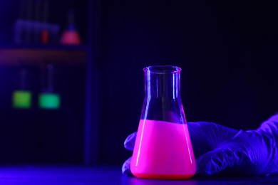Photo of Scientist with laboratory flask of luminous liquid at table against dark background, closeup. Space for text