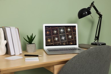 Photo of Modern laptop, books, lamp and stationery on wooden desk near green wall. Home office