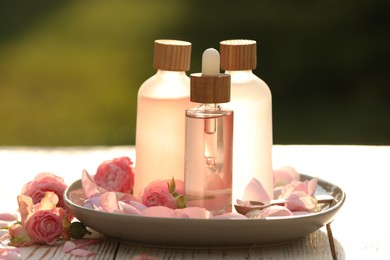 Photo of Bottles of rose essential oil and flowers on wooden table outdoors