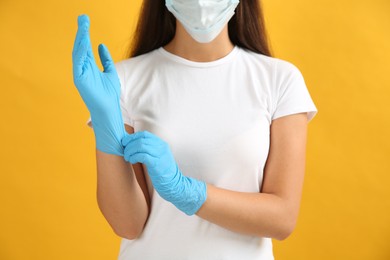Photo of Woman in protective face mask putting on medical gloves against yellow background, closeup