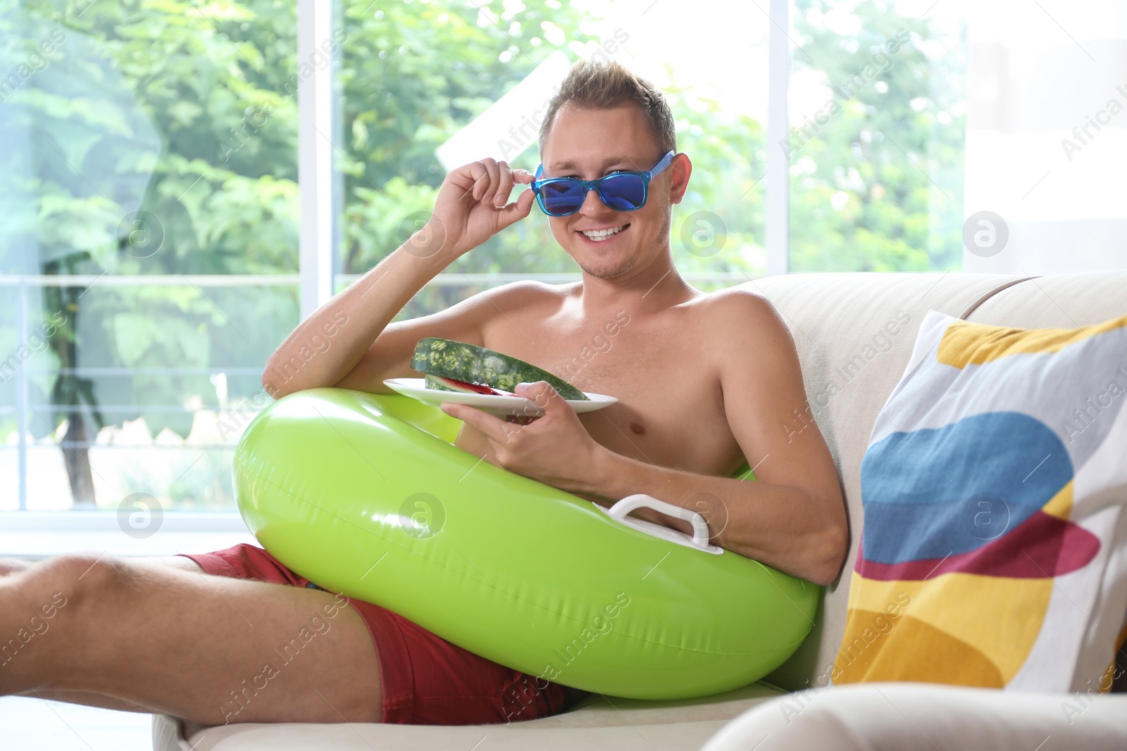 Photo of Shirtless man with inflatable ring and watermelon on sofa at home