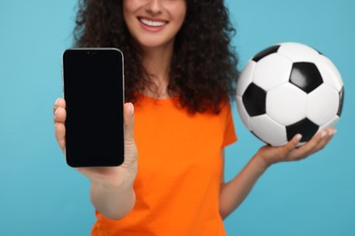 Happy fan holding soccer ball and showing smartphone on light blue background, closeup