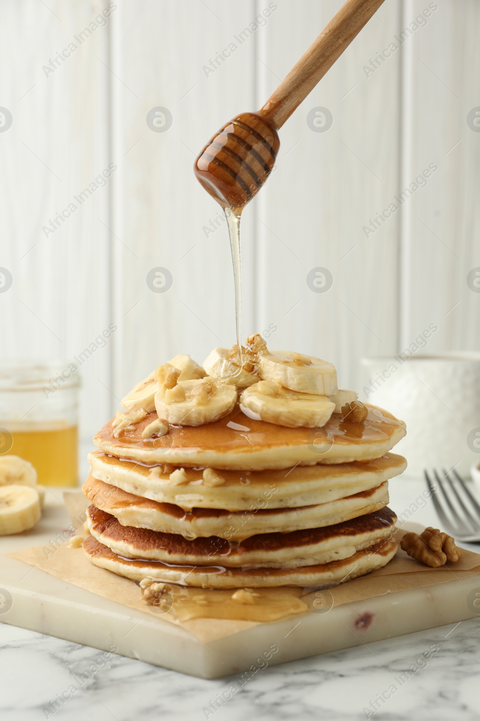 Photo of Pouring honey from dipper onto delicious pancakes with bananas and walnuts at white marble table