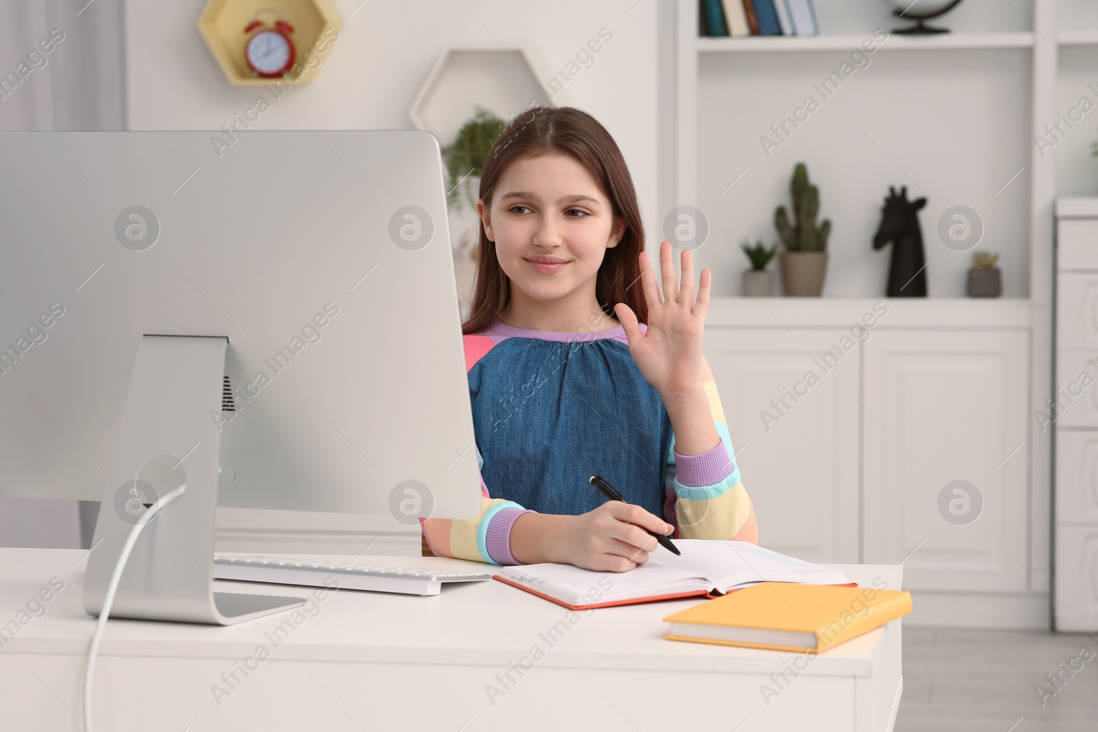 Photo of Cute girl writing in notepad while using computer at desk in room. Home workplace