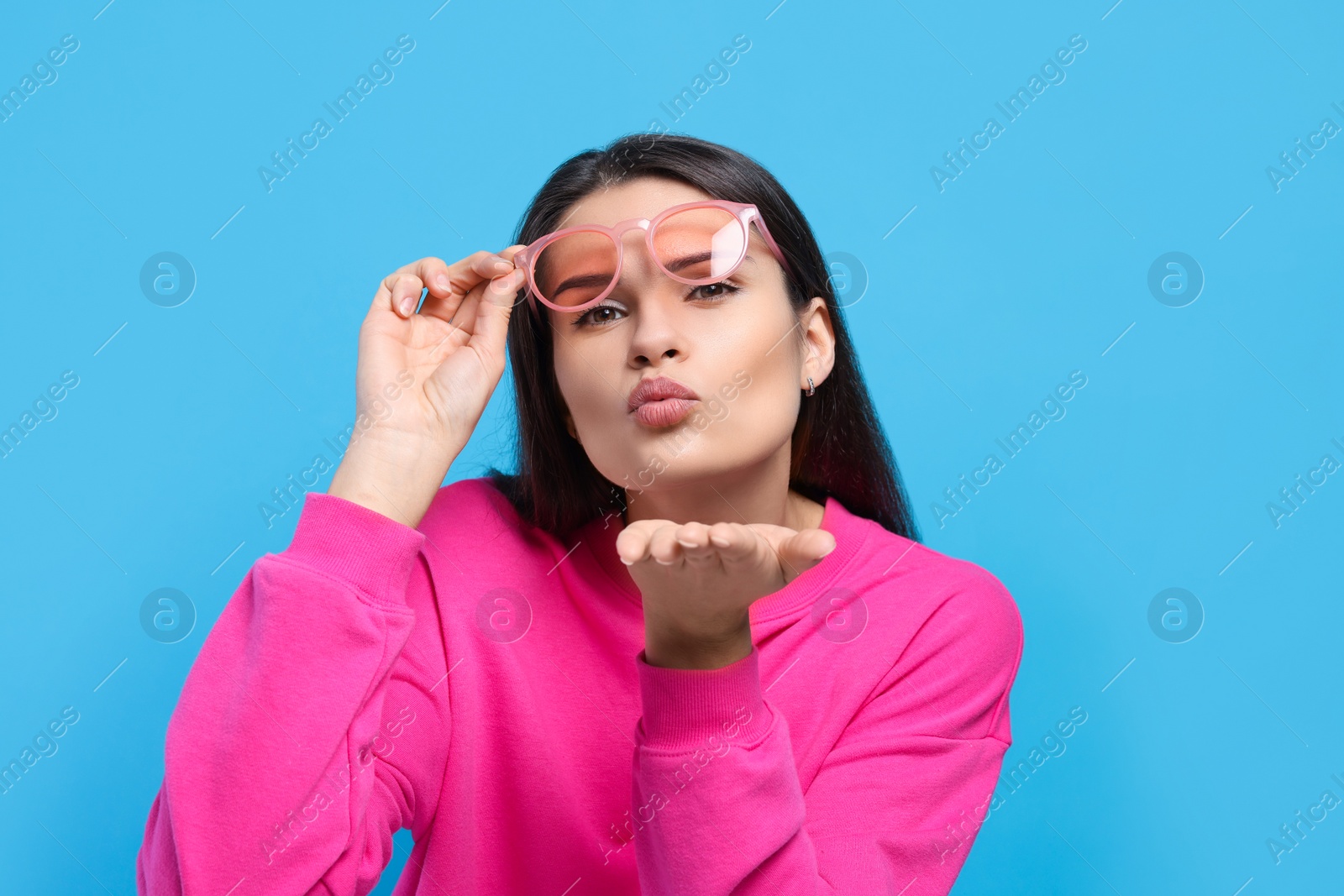 Photo of Beautiful young woman in stylish sunglasses blowing kiss on light blue background