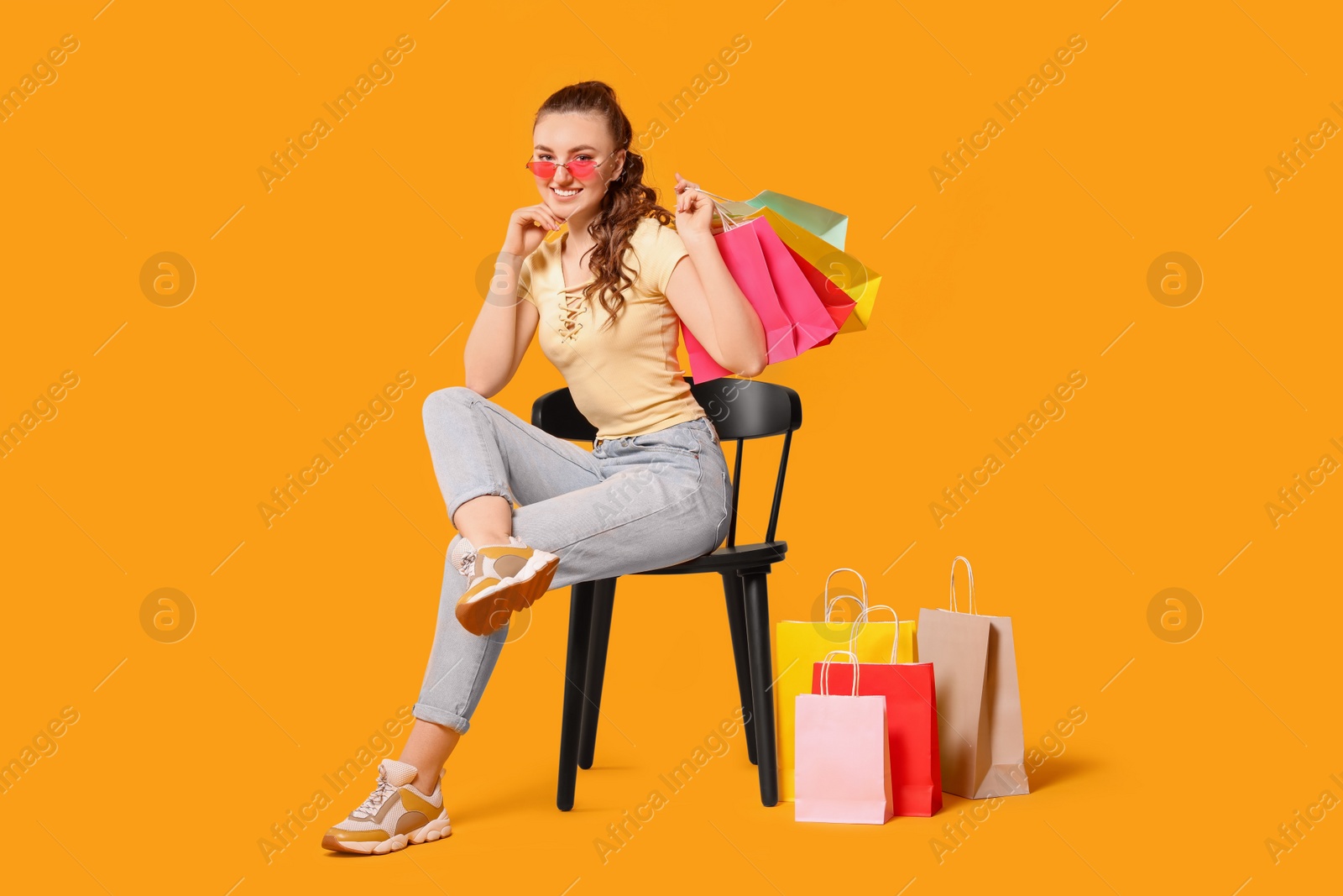 Photo of Happy woman in stylish sunglasses holding colorful shopping bags on chair against orange background