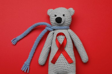 Photo of Cute knitted toy bear with ribbon on red background, top view. AIDS disease awareness