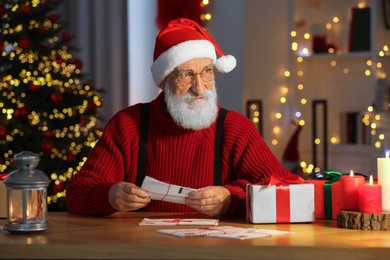 Santa Claus reading letter at his workplace in room decorated for Christmas