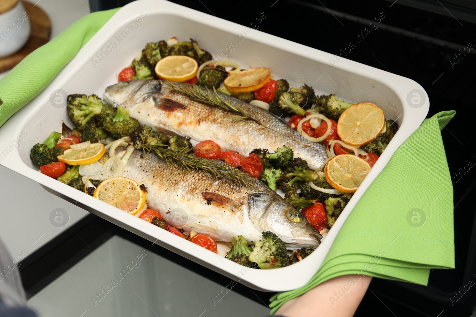 Photo of Woman taking baking dish with delicious fish and vegetables from oven in kitchen, closeup