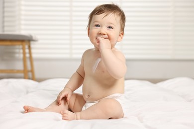 Photo of Cute little baby with moisturizing cream onto body sitting on bed at home