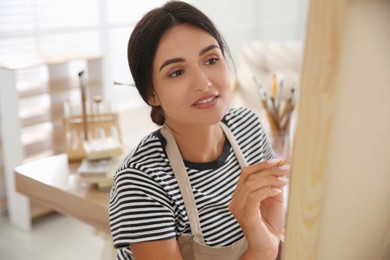 Photo of Young woman drawing on canvas with brush indoors