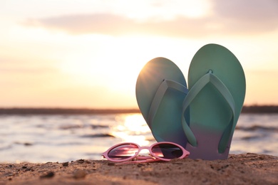 Stylish flip flops with sunglasses on sand near sea, space for text. Beach accessories