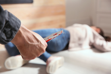 Photo of Man with bloody knife and his victim on floor indoors, closeup. Dangerous criminal