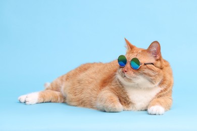 Photo of Cute ginger cat in stylish sunglasses lying on light blue background