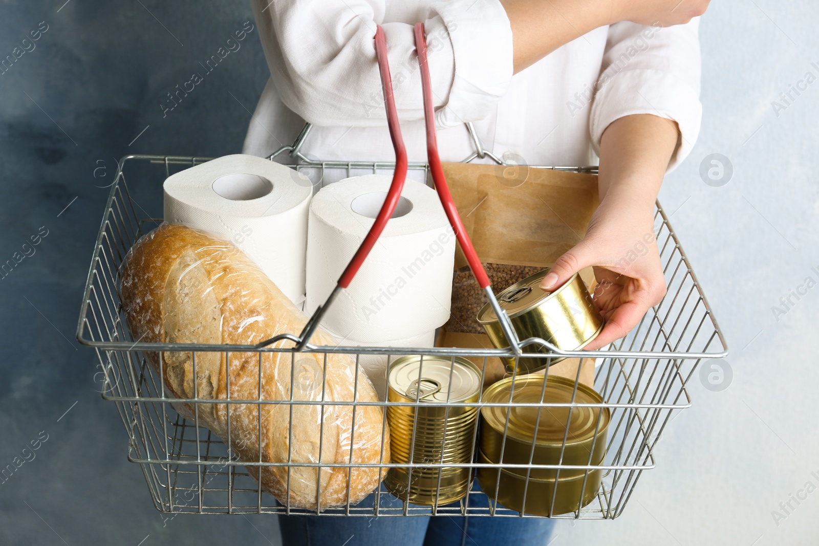 Photo of Woman holding shopping basket with products and toilet paper rolls on light blue background, closeup. Panic caused by virus