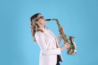 Beautiful young woman in elegant outfit playing saxophone on light blue background