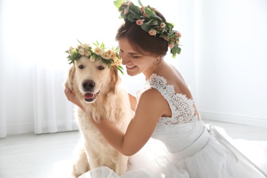 Photo of Bride and adorable Golden Retriever wearing wreath made of beautiful flowers indoors