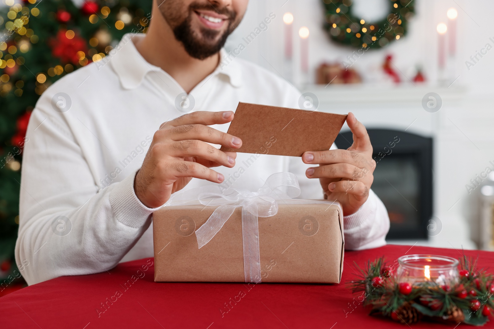 Photo of Man with Christmas gift reading greeting card at table indoors, closeup