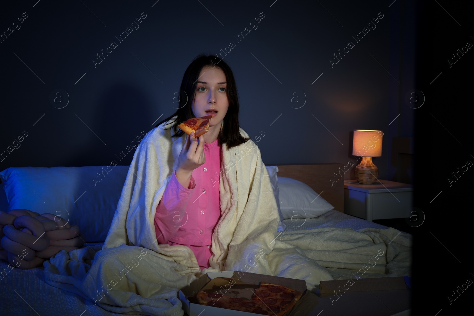 Photo of Young woman eating pizza while watching TV on bed at night