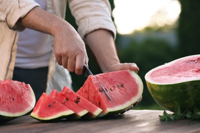 Photo of Man cutting tasty ripe watermelon at wooden table outdoors, closeup