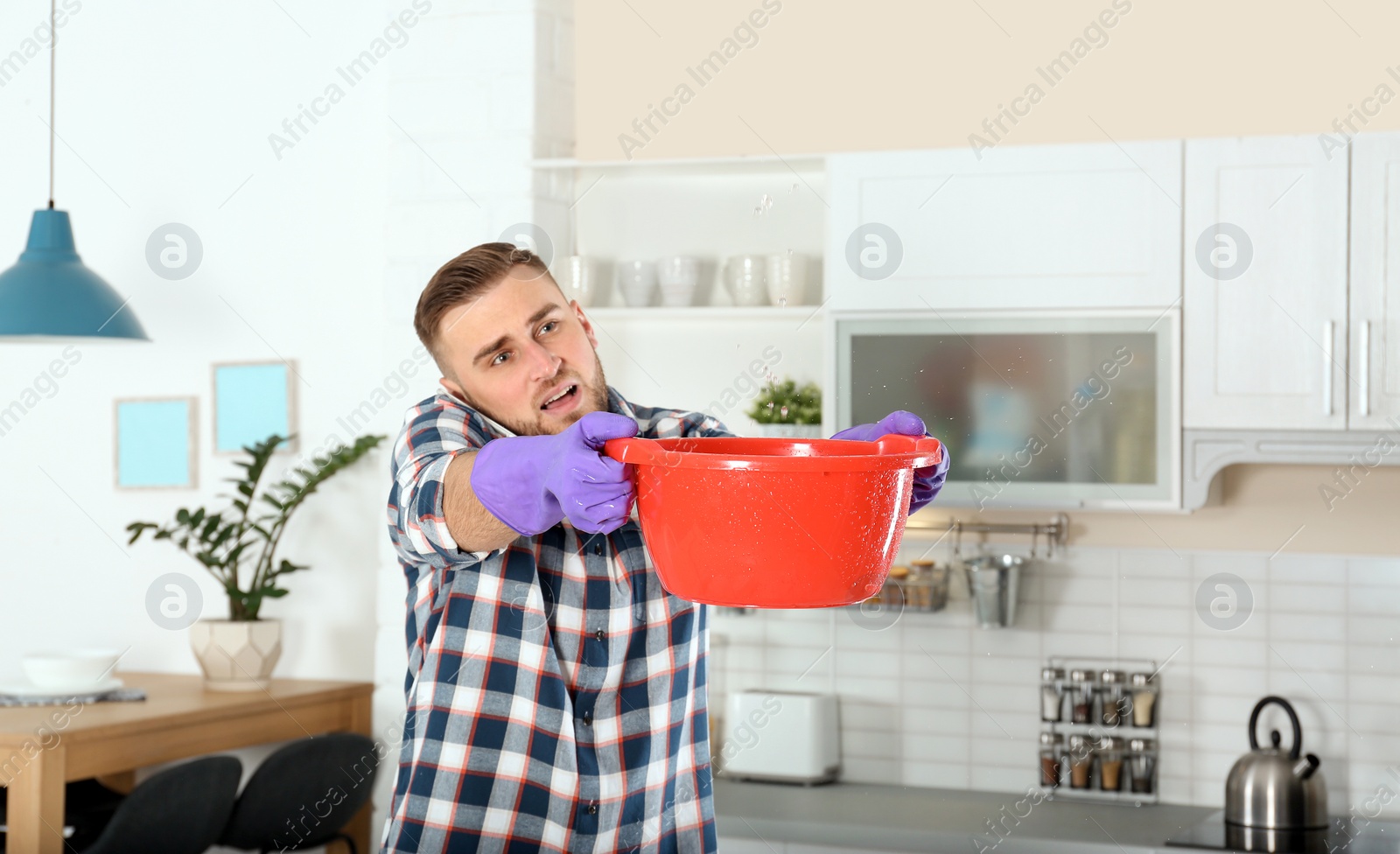 Photo of Emotional young man calling plumber while collecting water leakage from ceiling in kitchen