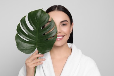 Photo of Woman in bathrobe holding leaf of monstera on light grey background. Spa treatment