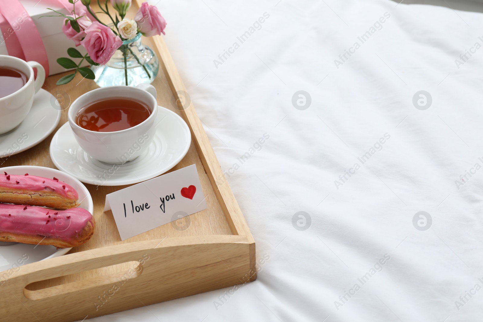 Photo of Tasty breakfast served in bed. Delicious eclairs, tea, gift box, flowers and card with phrase I Love You on tray, space for text