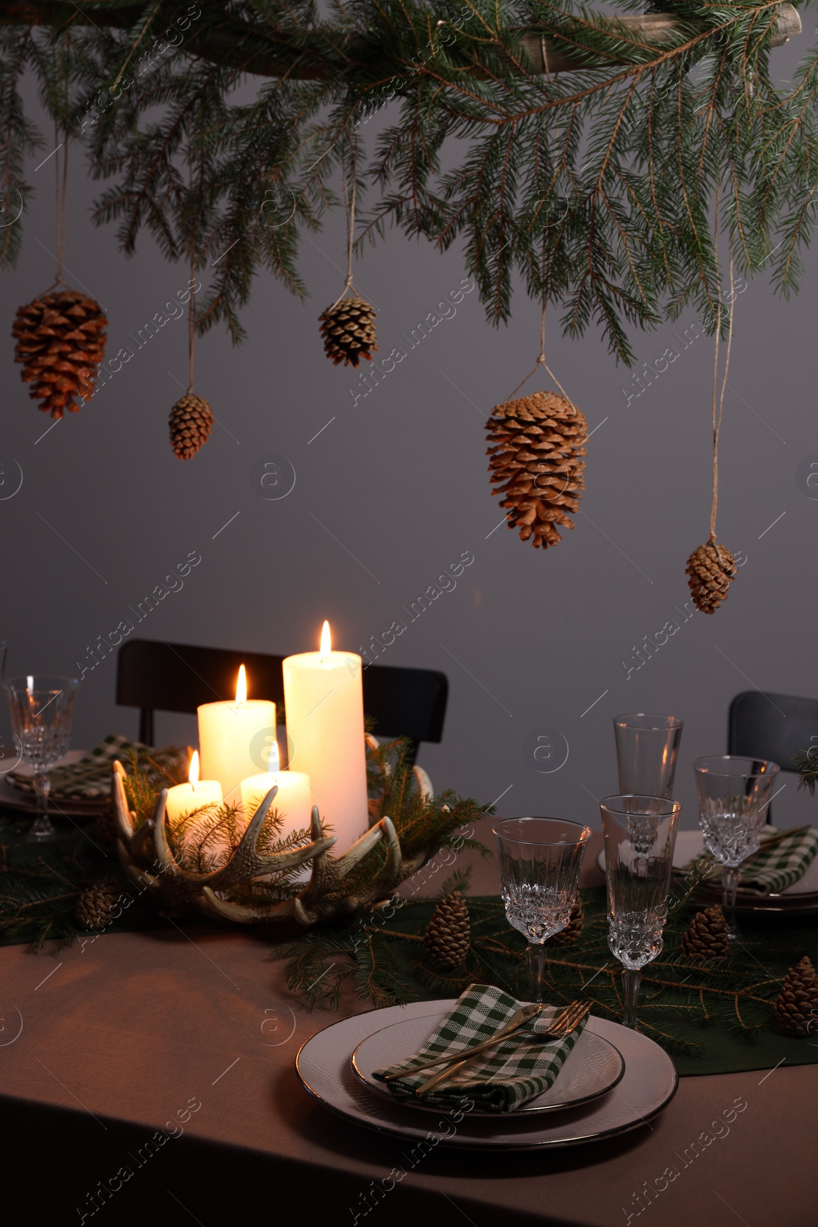 Photo of Christmas celebration. Cones hanging from fir tree branch over table with burning candles and tableware