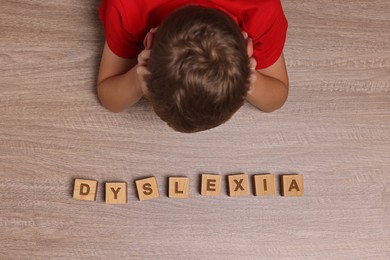 Little boy covering head with hands and cubes with word Dyslexia at wooden table, top view
