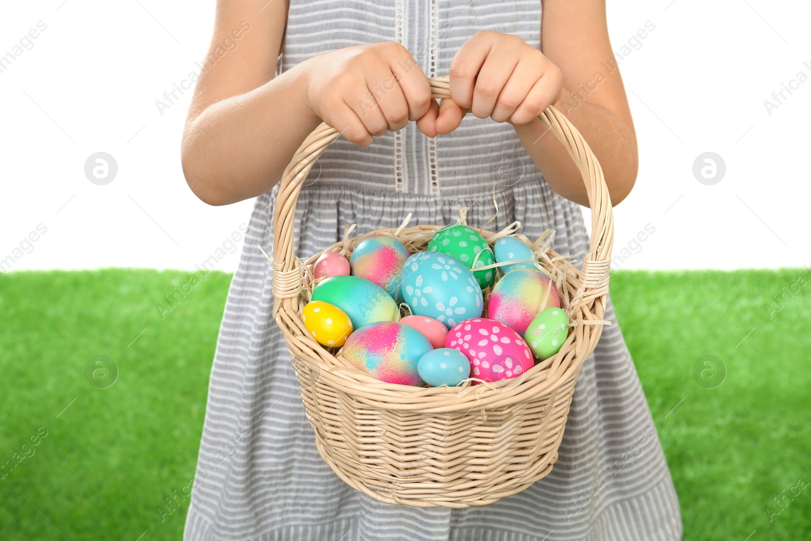 Photo of Little girl with basket full of Easter eggs on green grass against white background, closeup